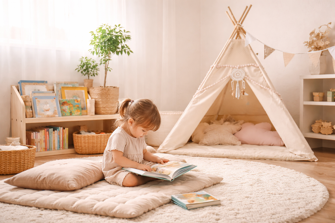 Kleines Kind sitzt auf einer weichen Matte im hellen Kinderzimmer und liest ein Bilderbuch; im Hintergrund steht ein beiges Tipi mit Kissen, daneben ein niedriges Bücherregal – gemütliche Leseecke im Kinderzimmer.