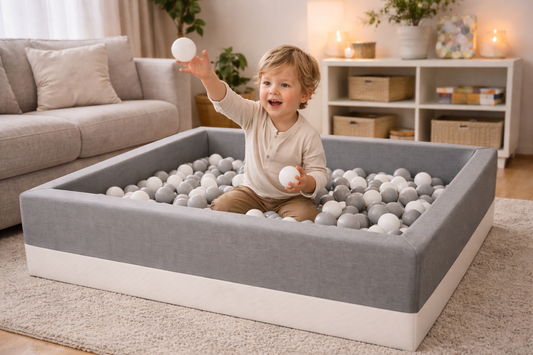 Dreijähriges Kind spielt fröhlich im grauen Bällebad im modernen Wohnzimmer mit weißen und grauen Bällen